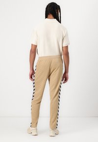 Punutudega juustega inimene, kes kannab valget lühikeste varrukatega särki, beežikaid dressipükse mereväesiniste V-kujuliste küljeribadega ja beeže tossusid, seisab seljaga vaataja poole.