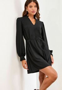 Schwarzes Kleid mit V-Ausschnitt, langen Ärmeln mit gepufften Schultern und Rüschensäumen. Mit einem Bindegürtel zur Anpassung und einer glatten Stofftextur.