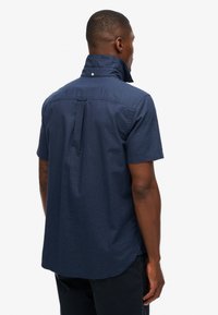 Camisa de manga corta en color azul marino con un cuello alzado, costuras visibles y un yugo trasero. La tela tiene una textura sutil. Dobladillo redondeado.