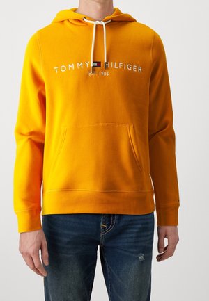 Mand iført en lyseorange hættetrøje med hvide snore og en forlomme, kombineret med mørkeblå jeans.