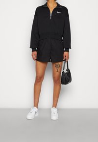 Person som bär en svart Nike zip-up sweatshirt, svarta shorts, vita Nike sneakers, håller en liten svart handväska och visar en blommig tatuering på låret.