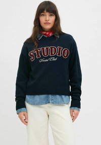 Jūras zils adīts džemperis ar uzrakstu "STUDIO" sarkanā krāsā. Iekļauts ribotās aproces un apakšmalas, uzvilkts virs gaiši zila džinsa krekla, apvienots ar baltām biksēm.