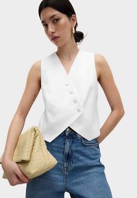 Weißes ärmelloses Wickeloberteil mit V-Ausschnitt und fünf bezogenen Knöpfen, kombiniert mit hoch taillierter blauer Jeans und einer gewebten beigefarbenen Clutch.