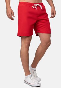 Pantaloni da bagno rossi con vita elastica e cordoncino bianco, caratterizzati da un design semplice senza fantasie. Indossati con sneakers bianche.