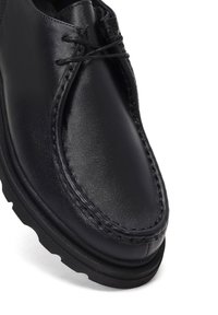 Chaussure en cuir noir de style mocassin avec bout arrondi, détails cousus, lacets noirs et semelle épaisse en caoutchouc.