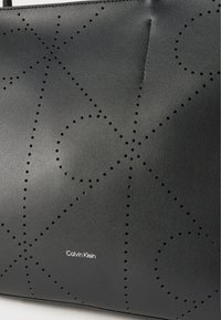 Μαύρη επιφάνεια από δέρμα με διάτρητο γεωμετρικό μοτίβο λογότυπου Calvin Klein και μικρό λευκό κείμενο με την επωνυμία Calvin Klein.