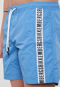 Costumi da bagno blu realizzati in tessuto leggero, con un cordoncino bianco e una striscia del logo nera lungo il lato. Texture liscia e leggermente lucida.