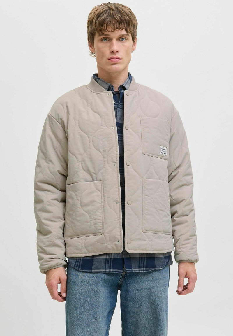Veste matelassée beige clair avec des boutons pression, dotée d'une poche avant et d'une finition texturée, portée par-dessus une chemise à carreaux sombre.