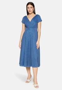 Femme portant une robe midi bleue pailletée à col en V avec manches courtes et des sandales compensées blanches, debout devant un fond blanc.