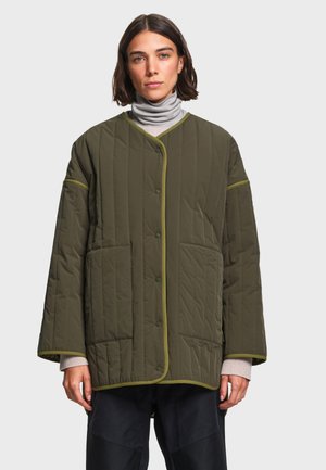Femme portant une veste matelassée vert olive oversize avec des boutons-pression, de grandes poches avant et un col roulé gris clair en dessous.