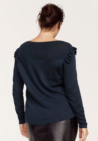 Navy modrý svetr s dlouhým rukávem, texturovaným horním zadním dílem a volánkovými akcenty na ramenních švech. Měkká tkanina s uvolněným střihem.
