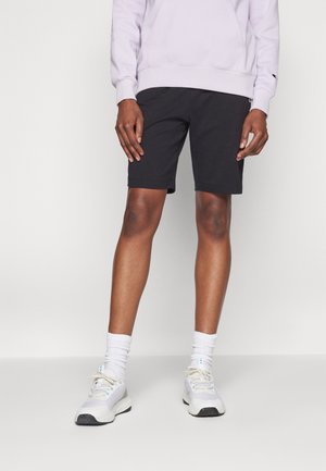 Persona vestida con pantalones cortos negros hasta la rodilla, zapatillas blancas, calcetas blancas hasta la pantorrilla y una sudadera color lavanda claro, de pie contra un fondo sencillo.