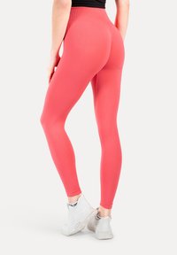 Vrouw die koraalrode leggings met hoge taille en witte sportschoenen draagt, staand met gekruiste benen tegen een effen achtergrond.