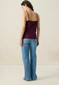 Vrouw in een mouwloze bordeauxrode top en lichtblauwe wijde jeans, staand met haar gezicht naar een beige muur en haar handen ontspannen aan haar zij.