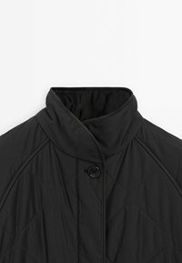 Veste matelassée noire avec col montant haut et fermeture à un bouton visible au niveau du cou sur un fond clair.