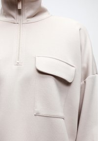 Ανοιχτό μπεζ πουλόβερ με φερμουάρ μέχρι το στήθος και μεγάλη τσέπη με καπάκι στο αριστερό στήθος, κατασκευασμένο από απαλό υλικό με υφή.