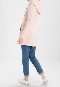 Abrigo rosa con capucha, tejido suave, diseño ajustado, longitud hasta el muslo, con bolsillos laterales. Combinado con jeans azul de puños enrollados y zapatos blancos.