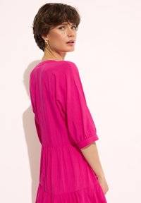 Fuksjowa sukienka wykonana z miękkiego materiału, z krótkimi bufiastymi rękawami, zebranym w talii i zaprojektowana w warstwowy sposób. Widoczne detale szwów.
