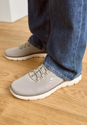 Zapatillas deportivas de malla gris con cordones elásticos y suela blanca, combinadas con jeans de mezclilla azul, de pie sobre un suelo de madera.
