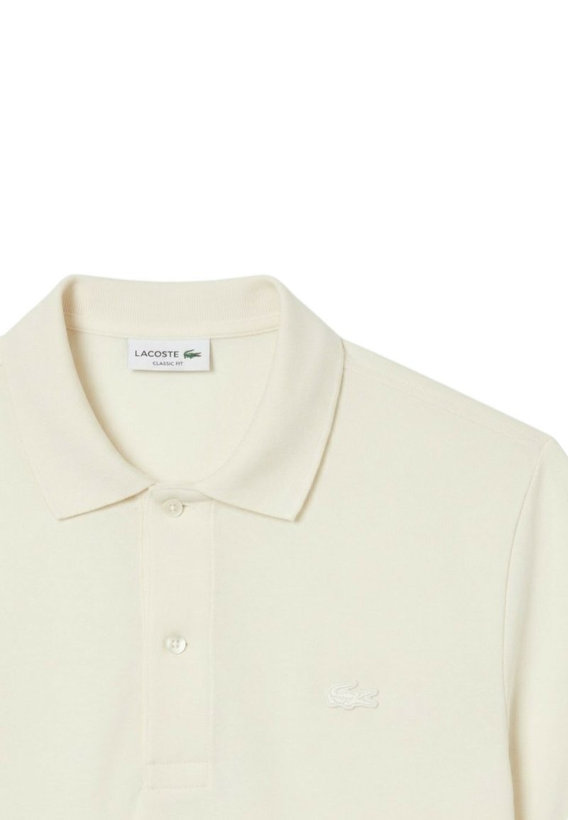 Roomkleurig Lacoste poloshirt in klassieke pasvorm met een kraag, twee knopen en een klein krokodillenlogo op de borst.