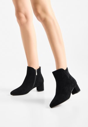Gambe snelle che indossano stivaletti neri in camoscio con zip laterale e bassi tacchi a blocchetto su sfondo bianco.