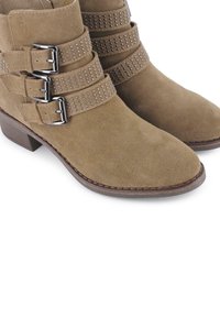 Botas de tobillo de ante marrón claras con tres correas ajustables con hebillas metálicas y acentos de tachuelas, tacón bajo apilado y punta afilada.