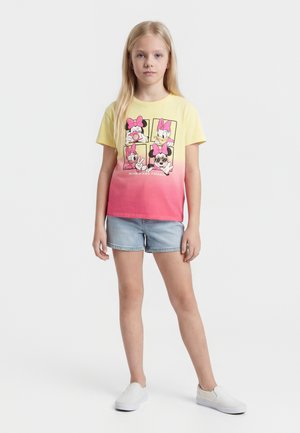 Blond dievča má na sebe žlto-ružové ombre tričko s grafikou Minnie Mouse, svetlé rifľové šortky a biele slip-on topánky, stojí na bielom pozadí.