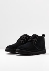 Des bottines noires en suede se caractérisent par un design à lacets, un bout arrondi et une semelle en caoutchouc texturée avec un accent de marque sur le talon.