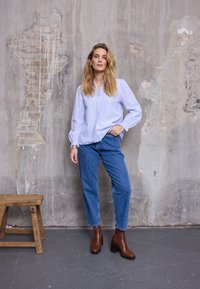 Blouse rayée bleue et blanche avec manches bouffantes, associée à un jean bleu taille haute et des bottines marron à talons. Fond neutre.