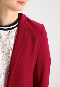 Femme portant un haut en dentelle blanche sous un blazer rouge avec un col rayé noir et blanc, cadrée au niveau du menton et de l'épaule.