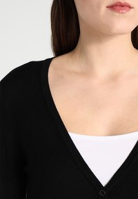 Svart v-ringad cardigan i mjukt tyg med knappstängning, bärs över en vit topp, vilket framhäver en slät textur och enkel design.