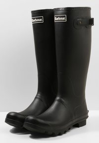 Botas de borracha à prova d'água, pretas, com uma textura suave, ponta arredondada e um sutil logótipo da marca em relevo. Apresenta uma correia na lateral para suporte.