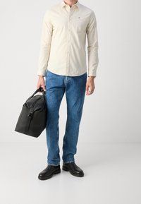 Camicia beige con bottoni, jeans in denim blu scuro, stivali di pelle nera e una borsa nera testurizzata con tracolla, dotata di chiusura zip superiore.