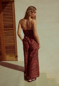 Röd och svart mönstrad byxdress med spagettiband, smockad bodice, vida ben och en texturerad tyg, bärs med platta sandaler.