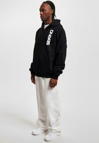 Haut à capuche zippé noir avec écriture blanche sur le côté gauche, associé à un pantalon blanc ample et des baskets blanches, sur un fond uni.