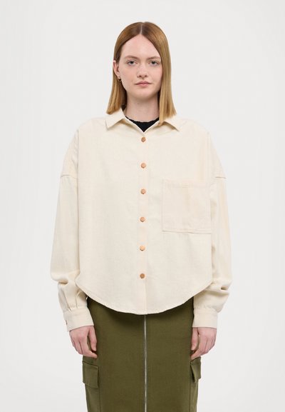 Camisa larga beige con botones, con un dobladillo redondeado, que presenta un solo bolsillo en el pecho y botones de cobre, llevada sobre una falda verde oliva.