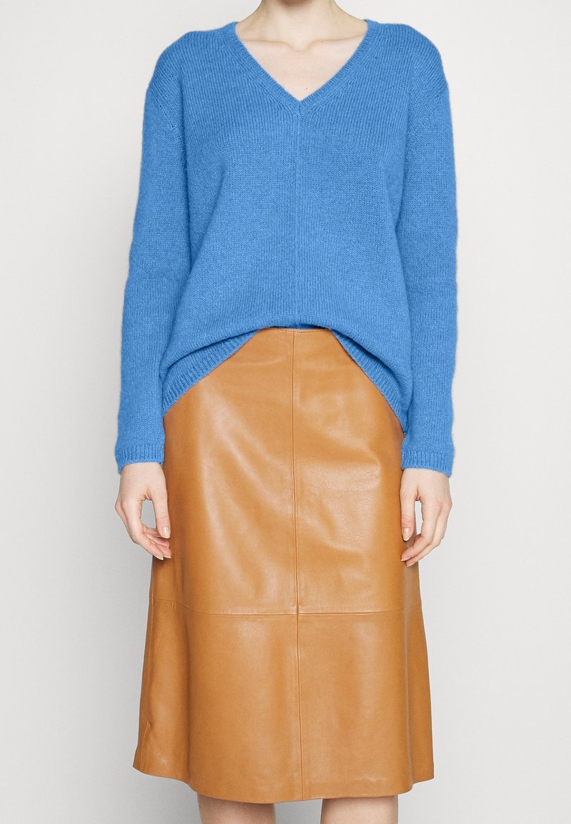 Femme portant un pull bleu col V rentré dans une jupe midi en cuir beige taille haute, debout devant un fond neutre.