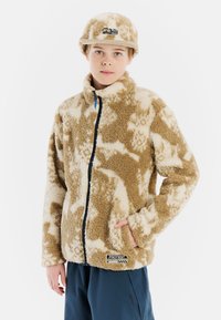 Chaqueta de forro polar beige con un patrón, cuello alto, cremallera frontal y bolsillos laterales; llevada con una gorra beige que presenta un logotipo en la parte delantera.