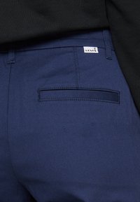 Pantaloni blu scuro con una trama liscia e una tasca posteriore, caratterizzati da un piccolo logo bianco Levi's cucito sopra la tasca.