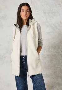 Lichtgrijze mouwloze fleece vest met capuchon, voorrits en twee zijzakken; gedragen over een grijze lange mouwen shirt en blauwe jeans.