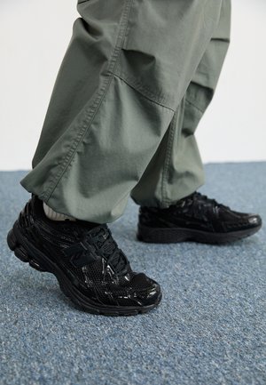 Personne portant un pantalon vert ample et des baskets noires brillantes, debout sur un sol recouvert d'un tapis bleu.