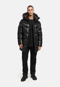 Svart puffjacka med en blank finish, utrustad med huva, dragkedjestängning, sidofickor och ett orange foder. Bärs tillsammans med svarta byxor och stövlar.