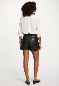 Pantaloni corti in pelle nera con tasche laterali, dettagli cuciti e una finitura liscia, abbinati a una camicia bianca con bottoni.