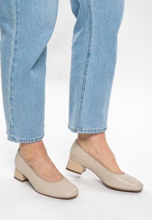 Beige schoenen met lage blokhiel en een gestructureerd patroon, gedragen met lichtblauwe cropped jeans op een witte achtergrond.