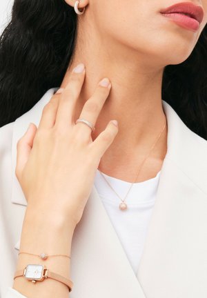 Orologio rosa oro con cinturino a rete, anelli scintillanti e collana con pendente caratterizzata da un dettaglio rotondo incastonato di gemme, abbinati a un blazer chiaro e a un top.