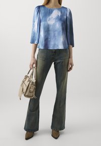 Blouse bleu tie-dye à manches trois quarts, assortie à un jean en denim bleu évasé. Un sac à main beige clair est tenu dans la main droite.