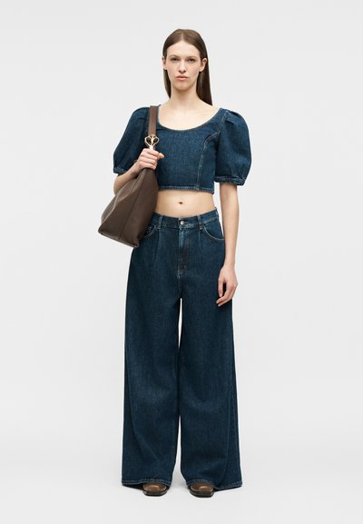 Femme portant un crop top en denim foncé à manches bouffantes et un jean à jambes larges, tenant un grand sac à bandoulière marron avec une fermeture dorée.
