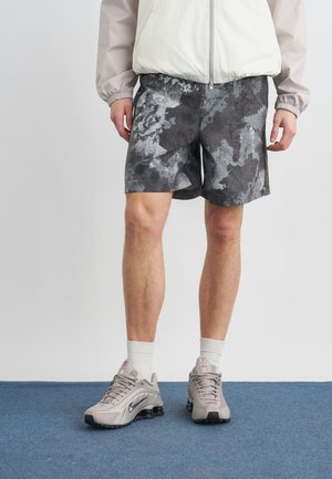Persoon die grijze en zwarte camouflage shorts draagt, een lichtgrijze windjack, witte sokken en beige Nike-sneakers, staand op een blauw tapijt.