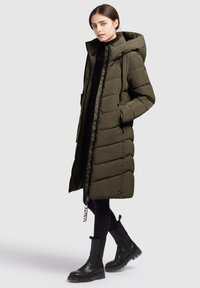 Olive zaļa pūkaina jaka ar kapuci, ar garu griezumu, šūnu dizainu un priekšējo rāvējslēdzēju. Valkāta kopā ar melnām legingām un masīvām melnām zābakiem.