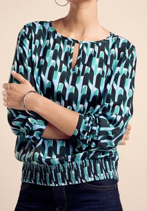 Vrouw draagt een blauw, zwart en wit patroon blouse met lange mouwen, een klein sleutelgat aan de voorkant en een elastische tailleband, armen gekruist.